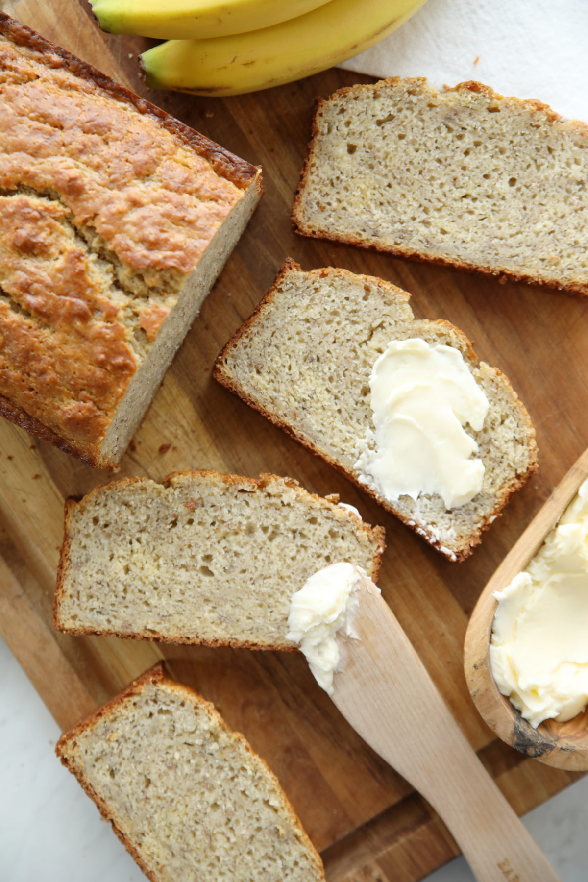 Sliced sourdough discard banana bread on a wood board with butter, a spreader, and fresh bananas
