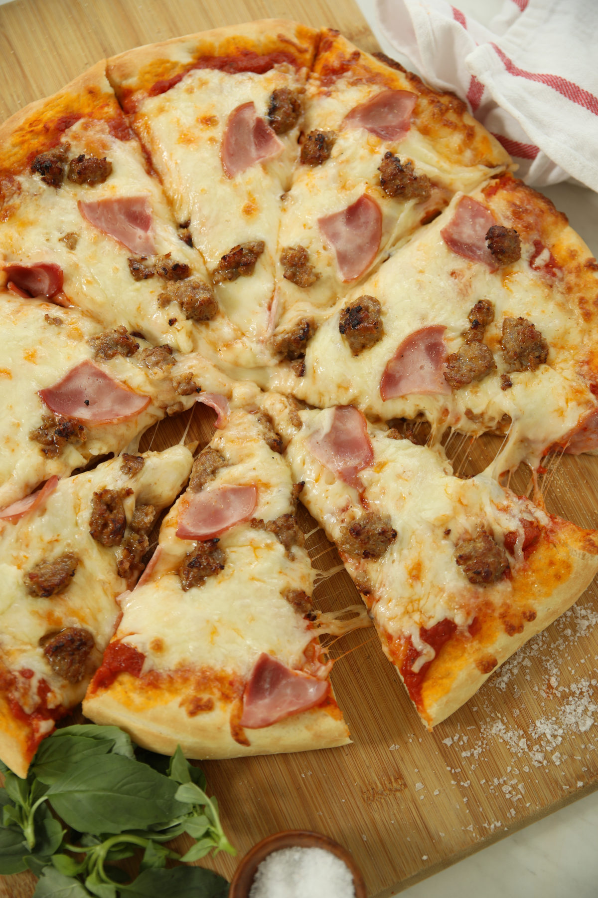 Overhead view of a sliced sourdough discard pizza with Italian sausage and Canadian bacon on a wooden cutting board with fresh basil and sea salt