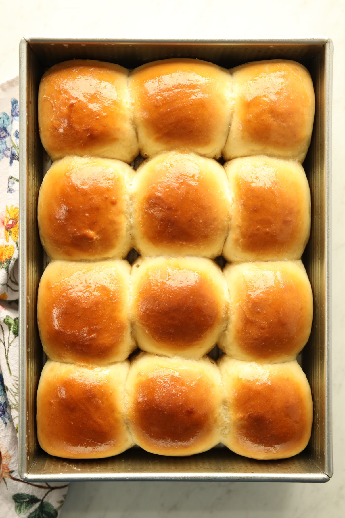 Twelve golden, butter-rubbed sourdough discard rolls fresh from the oven, filling a metal 9x13 baking pan with a floral tea towel alongside.