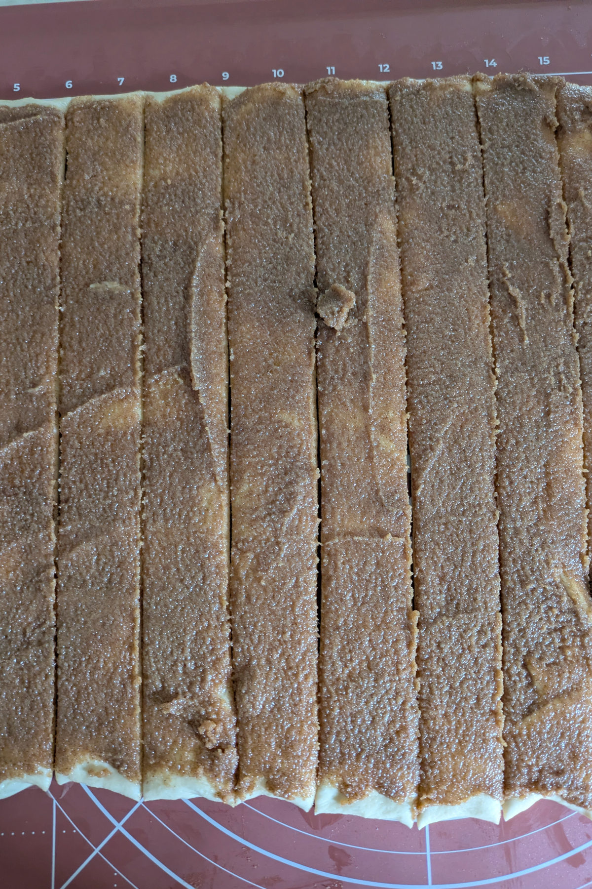 Sourdough cinnamon roll dough cut into strips with a pizza cutter on a silicone dough mat, showing the brown sugar cinnamon filling ready to be rolled.