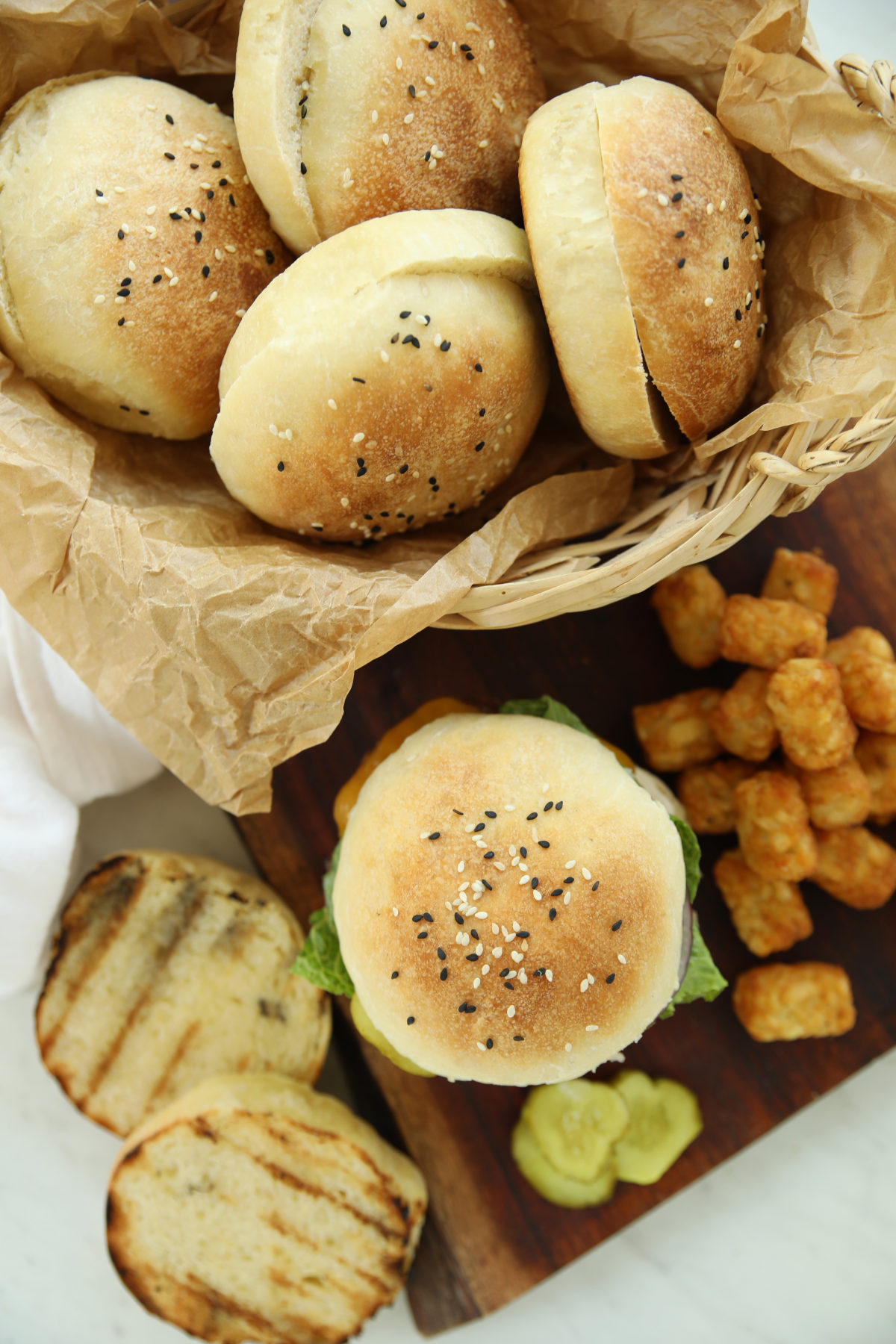 Overhead view of sourdough hamburger buns topped with black and white sesame seeds in a parchment-lined basket, with an assembled cheeseburger, toasted bun halves, tater tots, and pickle slices on a wooden board.