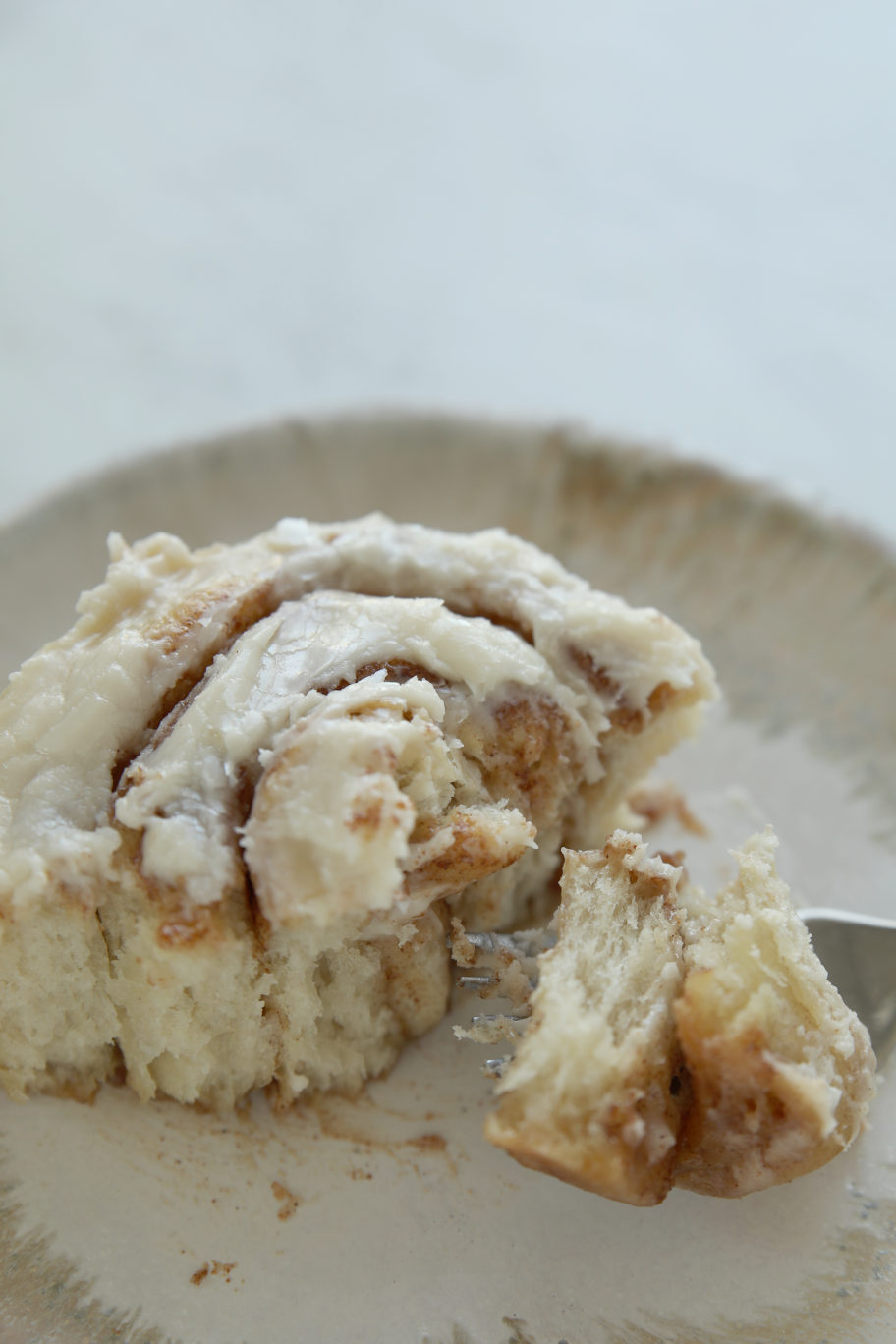 A fork pulling away a bite from a frosted sourdough cinnamon roll on a plate, showing the soft, fluffy interior layers and buttercream glaze.