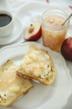 A jar of white nectarine freezer jam with a spoon resting in it, surrounded by fresh white nectarines and sliced toast on a white plate.