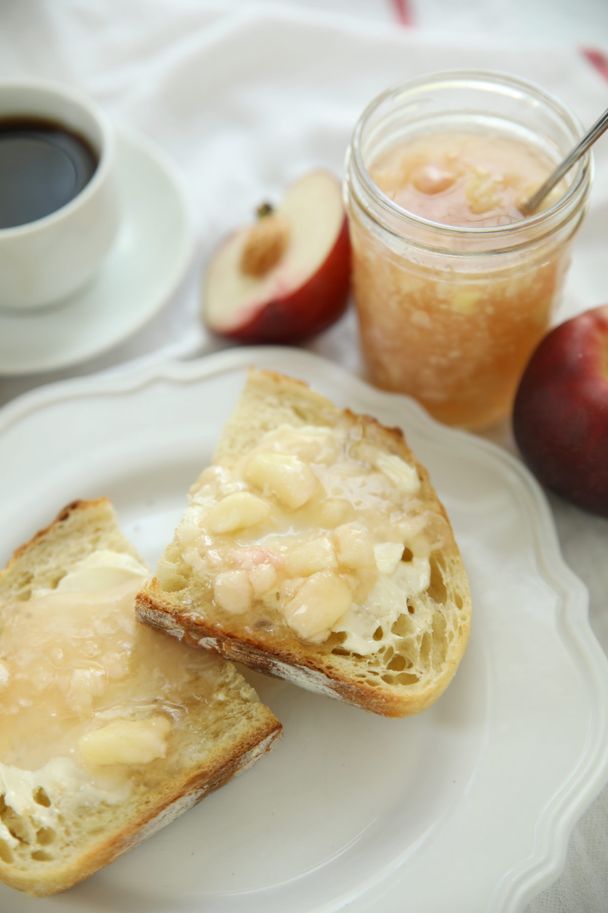 A jar of white nectarine freezer jam with a spoon resting in it, surrounded by fresh white nectarines and sliced toast on a white plate.