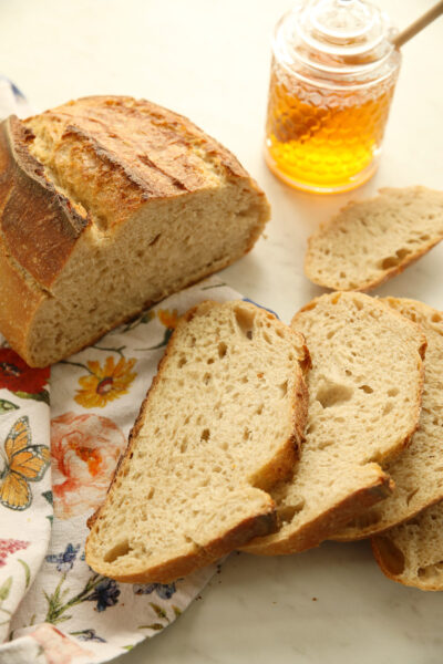 Sliced whole wheat sourdough bread on a floral cloth with a jar of honey, showing a golden crust and soft open crumb