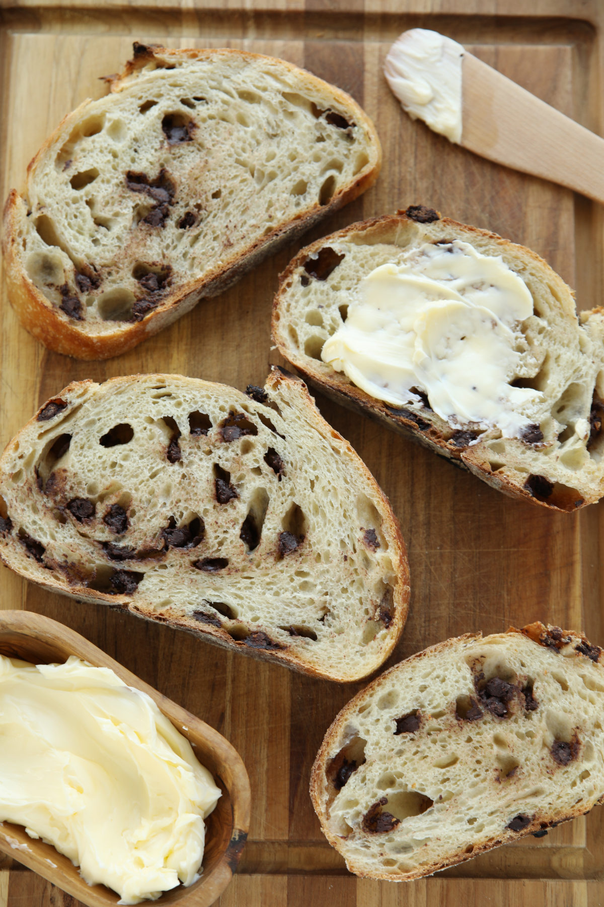 Sliced chocolate chip sourdough bread on a wood board with a buttered slice in front, showing the chocolate-flecked crumb