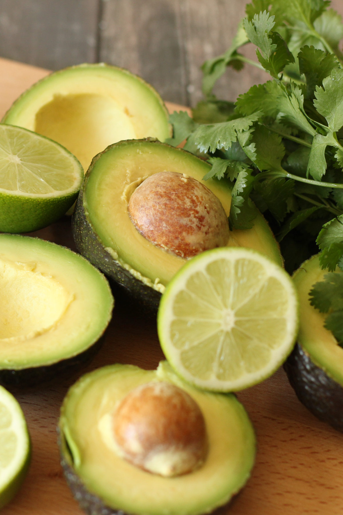 Halved ripe avocados with pits, lime halves, and fresh cilantro on a wood cutting board