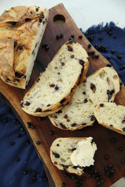 Sliced blueberry sourdough bread on a wood board with a buttered slice in front, showing the purple-flecked crumb