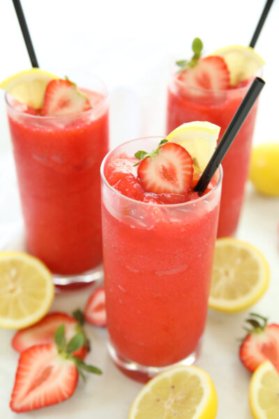 A tall glass of boozy frozen strawberry lemonade garnished with a strawberry fan and lemon wheel, with two more glasses and fresh lemons and strawberries in the background.
