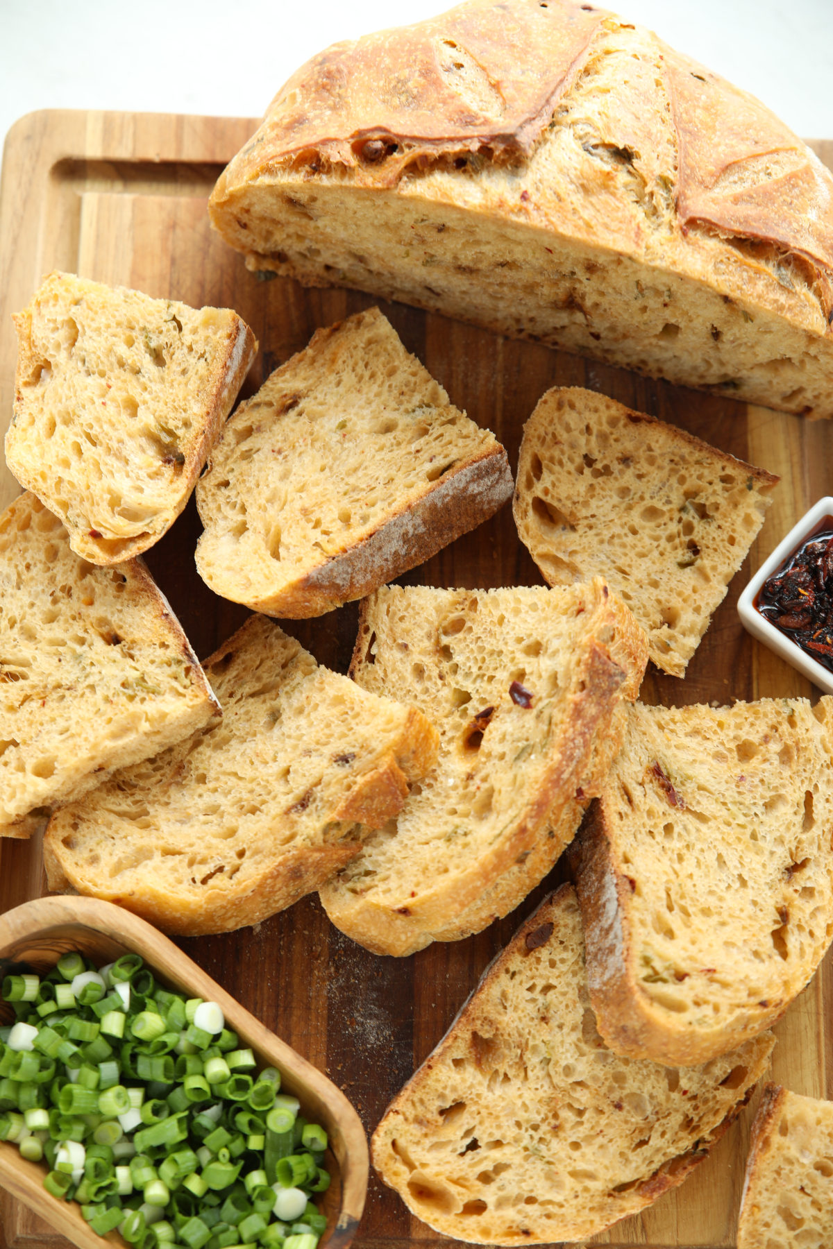 Sliced chili crisp green onion sourdough bread on a wood cutting board, with a bowl of chili crisp and chopped green onions alongside.