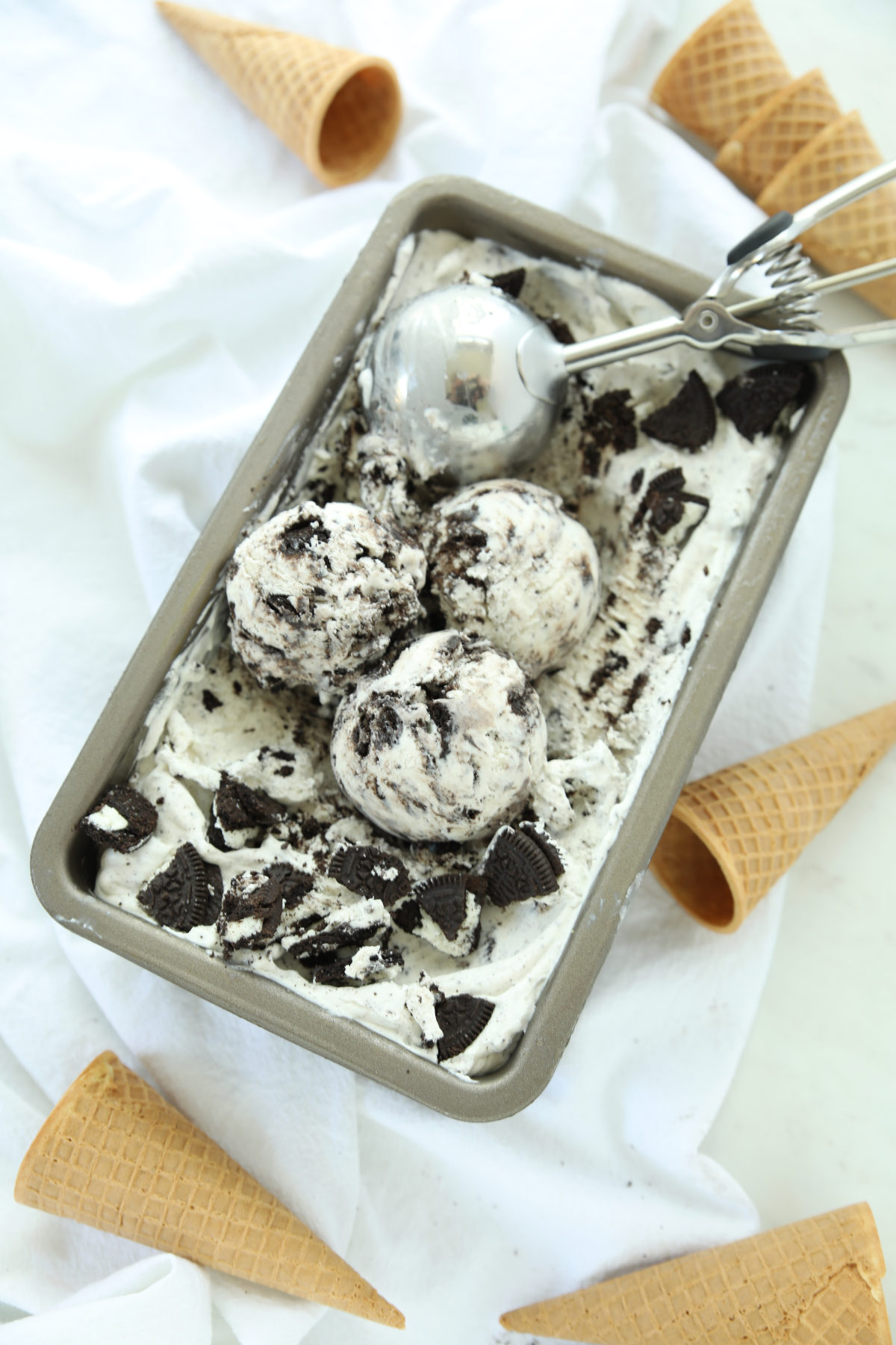 Three scoops of no-churn cookies and cream ice cream in a metal loaf pan, surrounded by waffle cones and crushed Oreo cookies