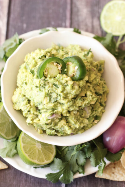 Chunky guacamole in a white bowl topped with jalapeño slices, surrounded by cilantro, lime wedges, red onion, and tortilla chips