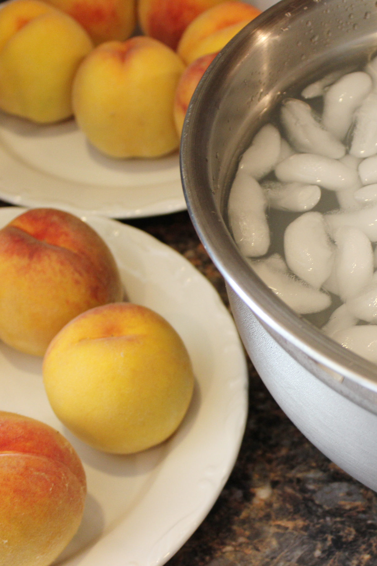 Fresh whole peaches on plates next to a bowl of ice water for blanching
