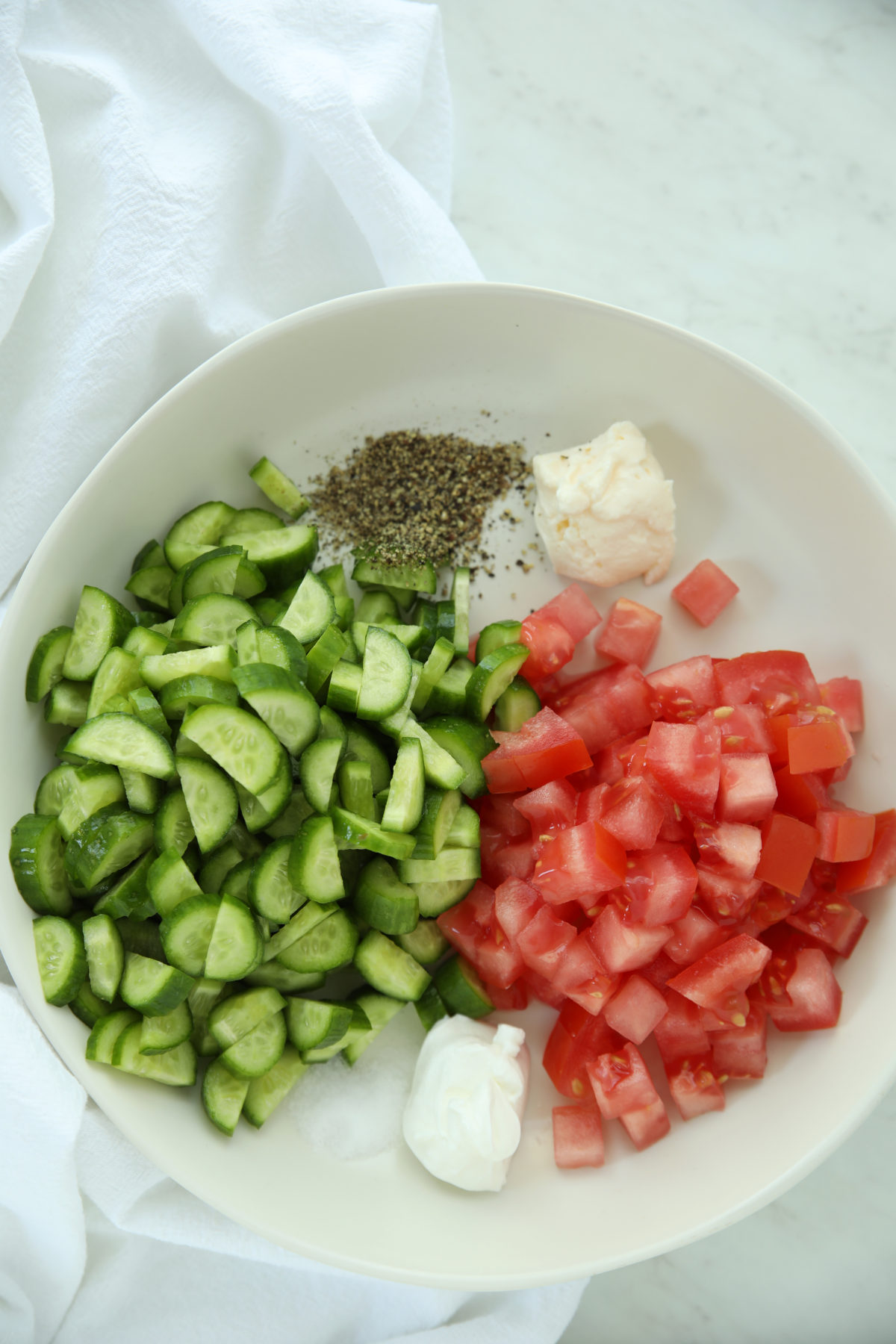Overhead view of a bowl with diced cucumbers and diced tomatoes for cucumber tomato salad, alongside mayonnaise, sour cream, and black pepper ready to be mixed together.