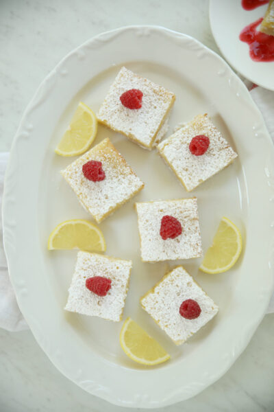 Overhead view of lemon bars with shortbread crust arranged on a white platter, each topped with powdered sugar and a fresh raspberry, with lemon wedges tucked between them.