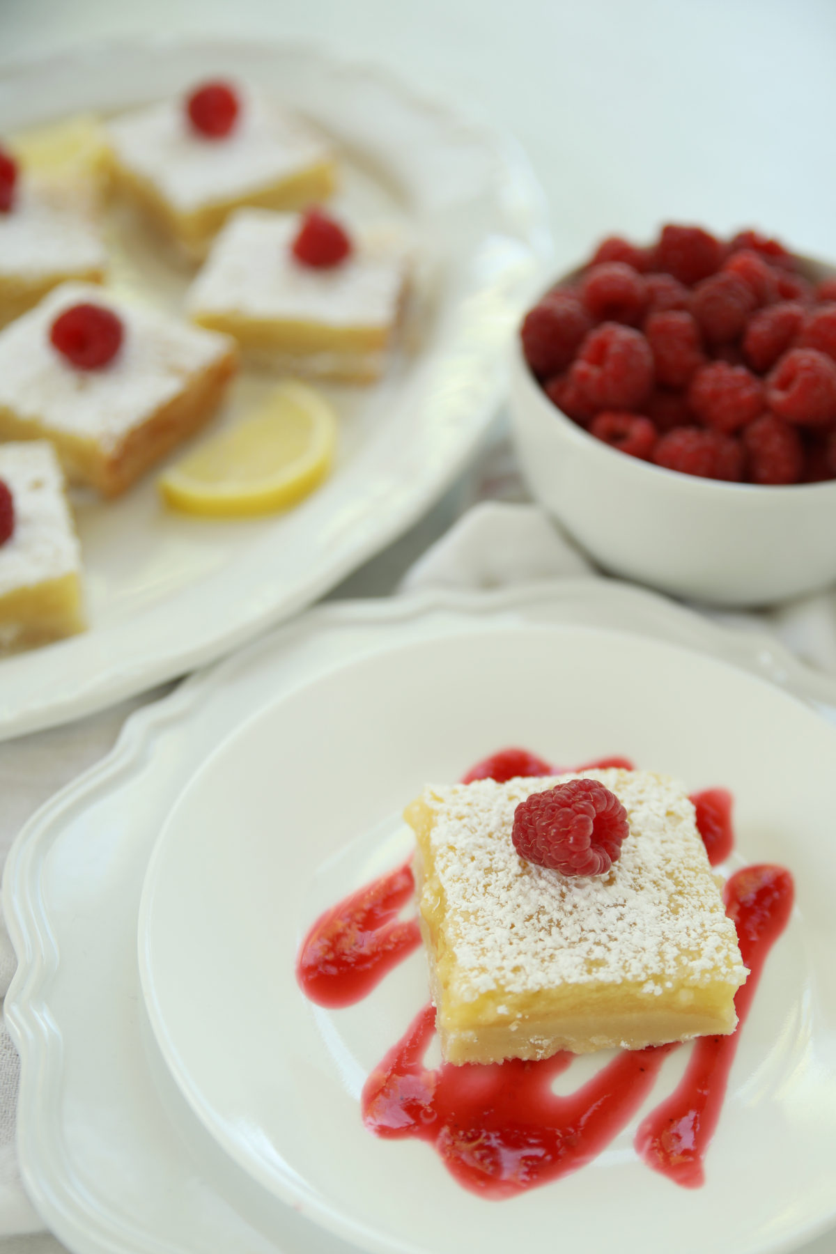 A single lemon bar with shortbread crust on a white plate, drizzled with raspberry sauce and topped with a fresh raspberry, with more bars and a bowl of raspberries blurred in the background.
