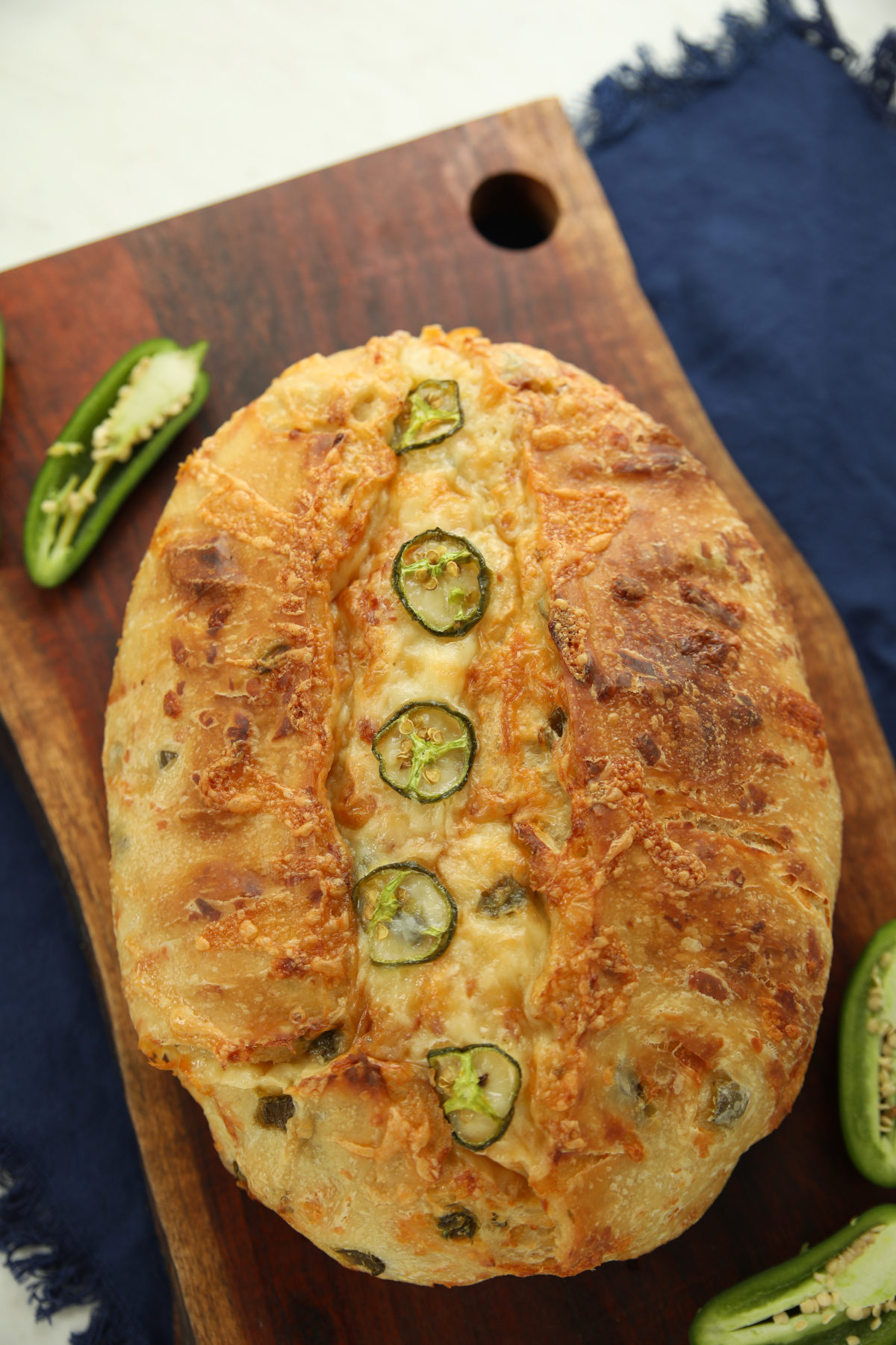 Overhead view of a whole jalapeño cheddar sourdough boule with a scored center, topped with melted cheese and jalapeño slices, on a wood board with fresh jalapeño halves alongside.
