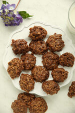 A pile of no bake chocolate oatmeal cookies on a white scalloped plate, with a purple hydrangea and a glass of milk alongside.