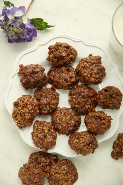 A pile of no bake chocolate oatmeal cookies on a white scalloped plate, with a purple hydrangea and a glass of milk alongside.