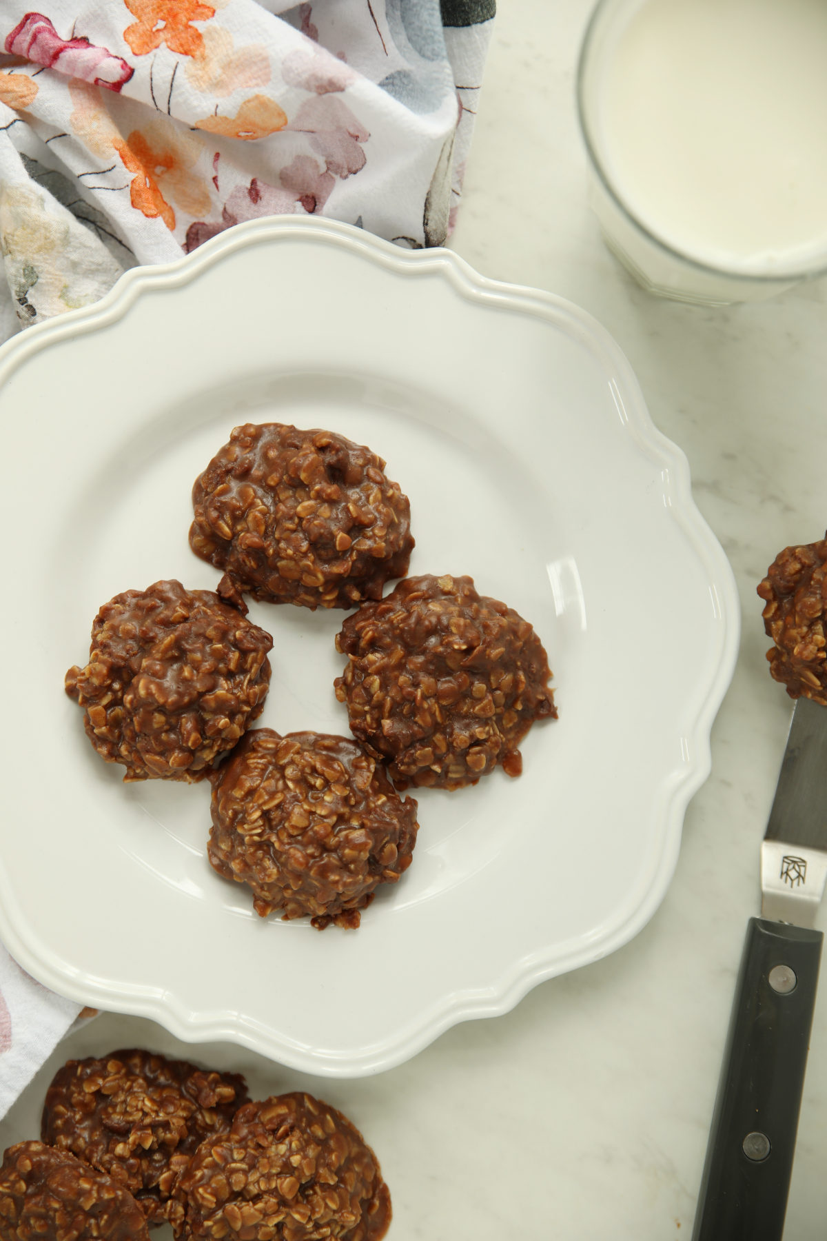 Four no bake chocolate oatmeal cookies arranged on a white scalloped plate, with a glass of milk, a knife, and a floral napkin nearby.