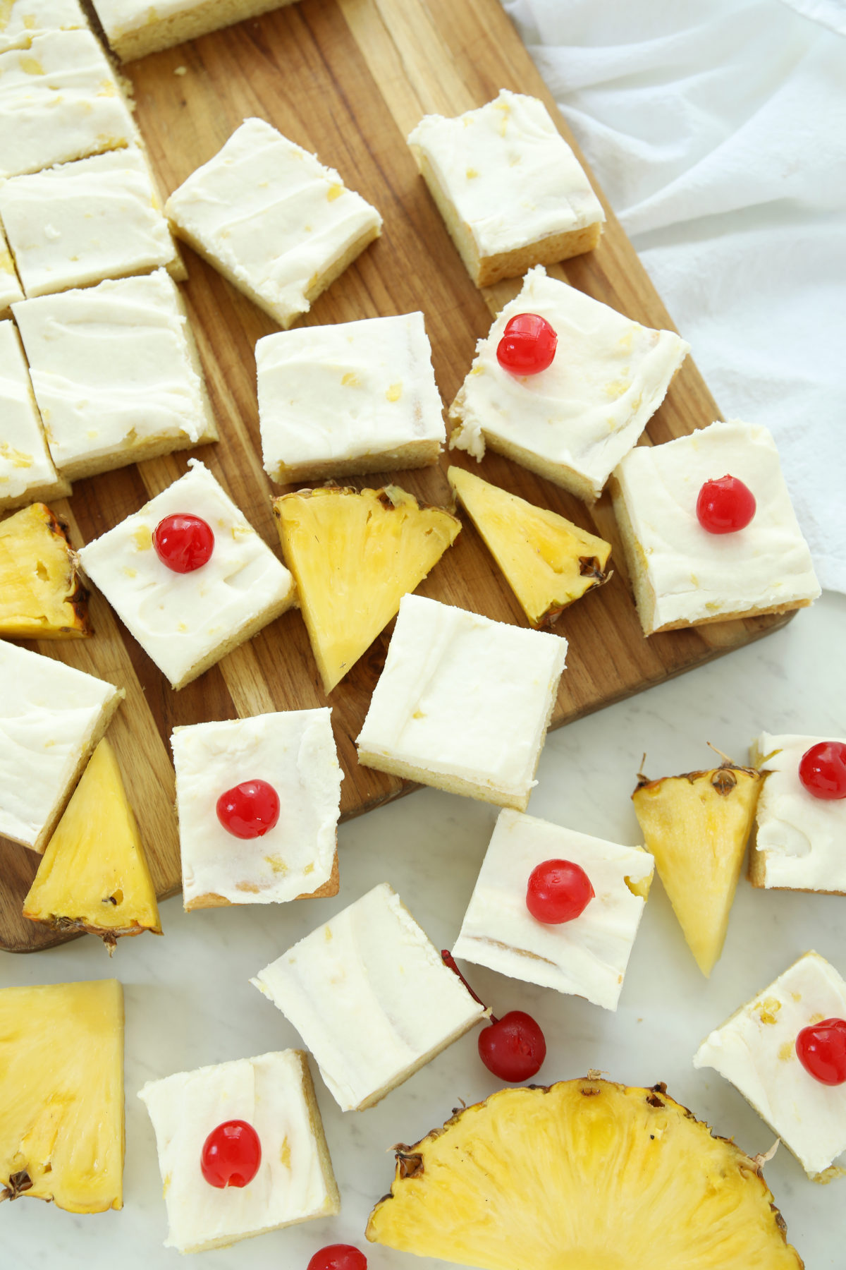 Pina colada cookie bars topped with coconut-pineapple buttercream and maraschino cherries, arranged on a wood cutting board with fresh pineapple wedges and a marble counter.