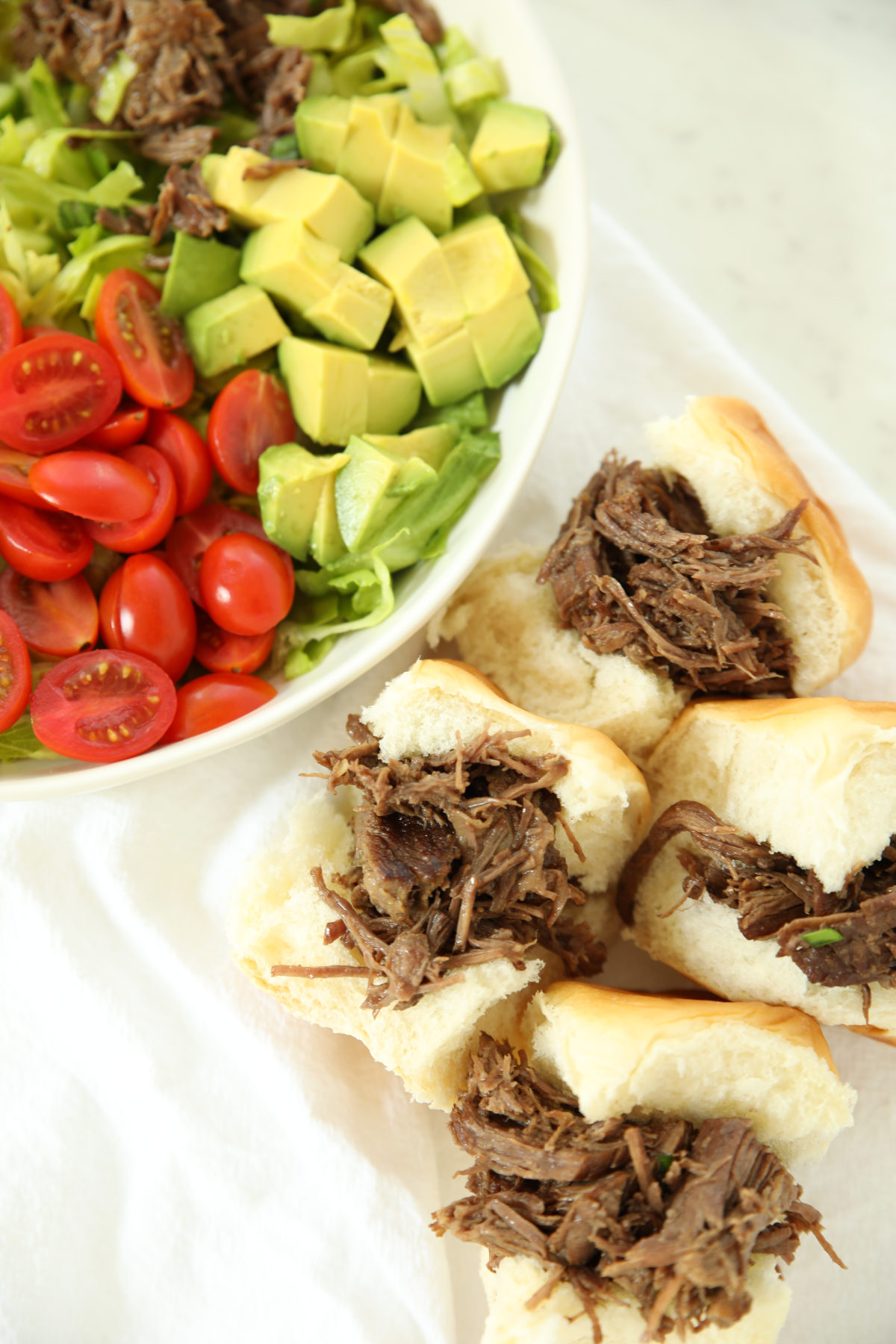 Pulled beef sliders on soft buns next to a bowl of salad with tomatoes and avocado