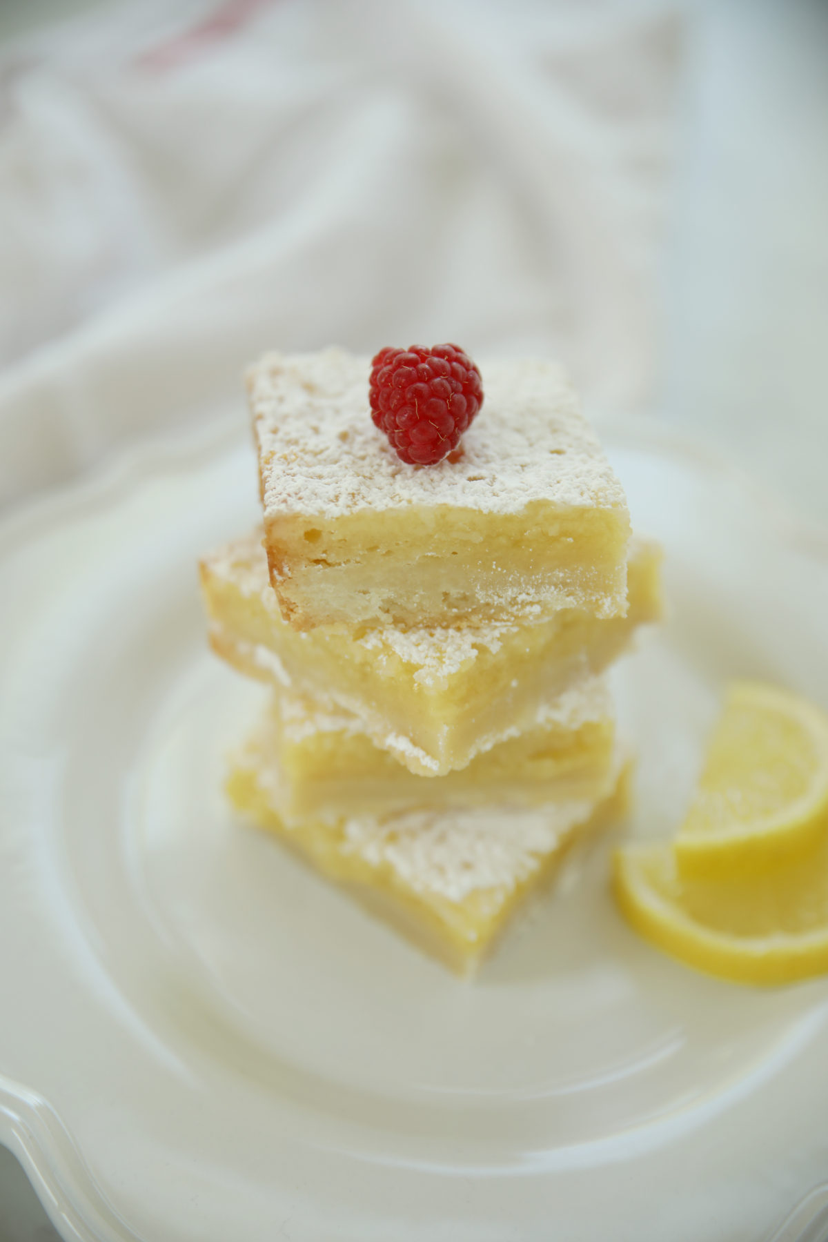 A stack of lemon bars with shortbread crust topped with a fresh raspberry and powdered sugar, next to a lemon wedge on a white plate.