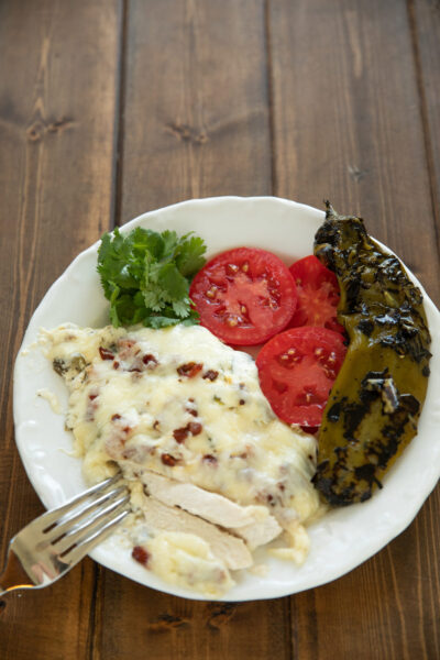 A white plate holds a serving of keto chicken casserole with melted cheese and crispy bacon, chicken sliced open on a fork, next to tomato slices, fresh cilantro, and a whole fire roasted hatch chile