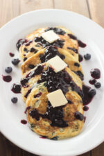 A white oval platter holds golden buttermilk blueberry pancakes topped with warm blueberry sauce and melting pats of butter, with fresh blueberries scattered around the plate on a wood table.