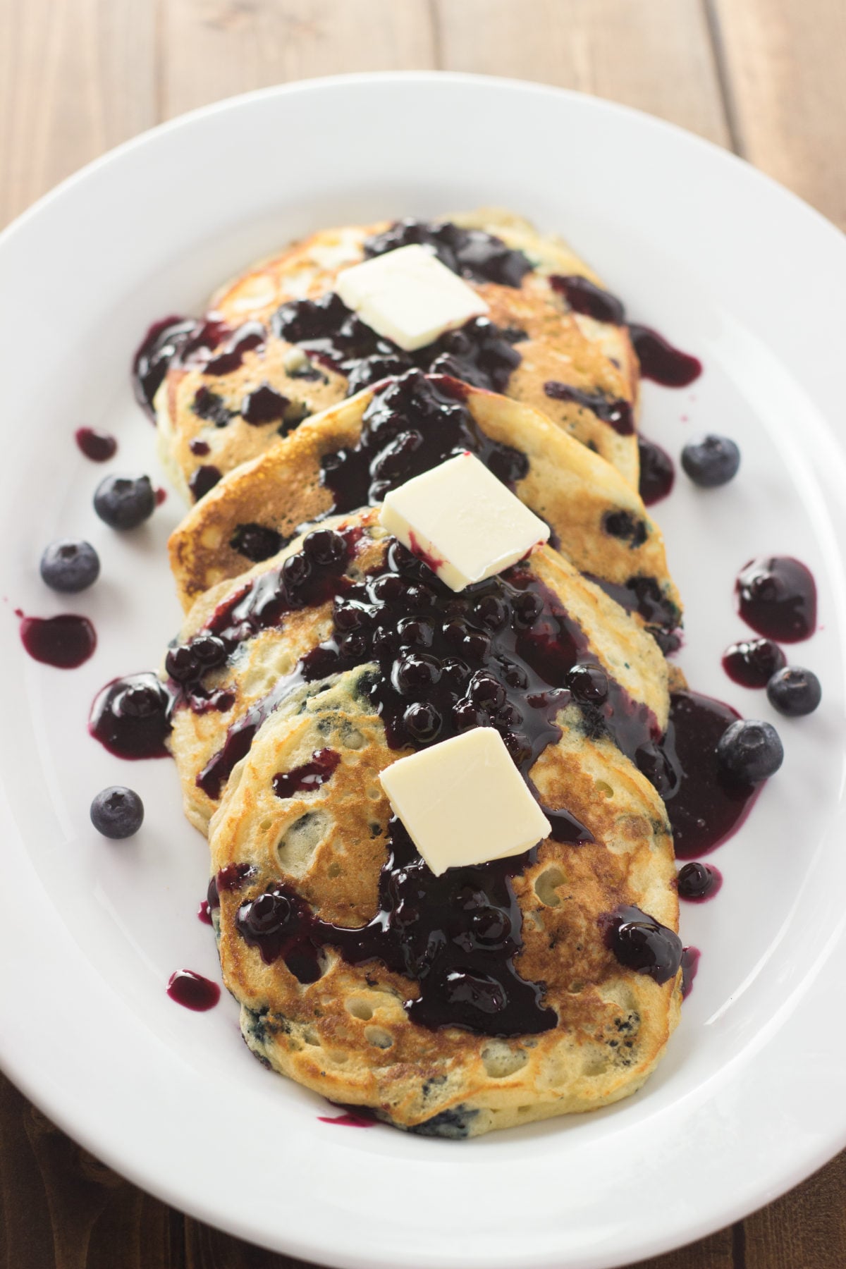 A white oval platter holds golden buttermilk blueberry pancakes topped with warm blueberry sauce and melting pats of butter, with fresh blueberries scattered around the plate on a wood table.