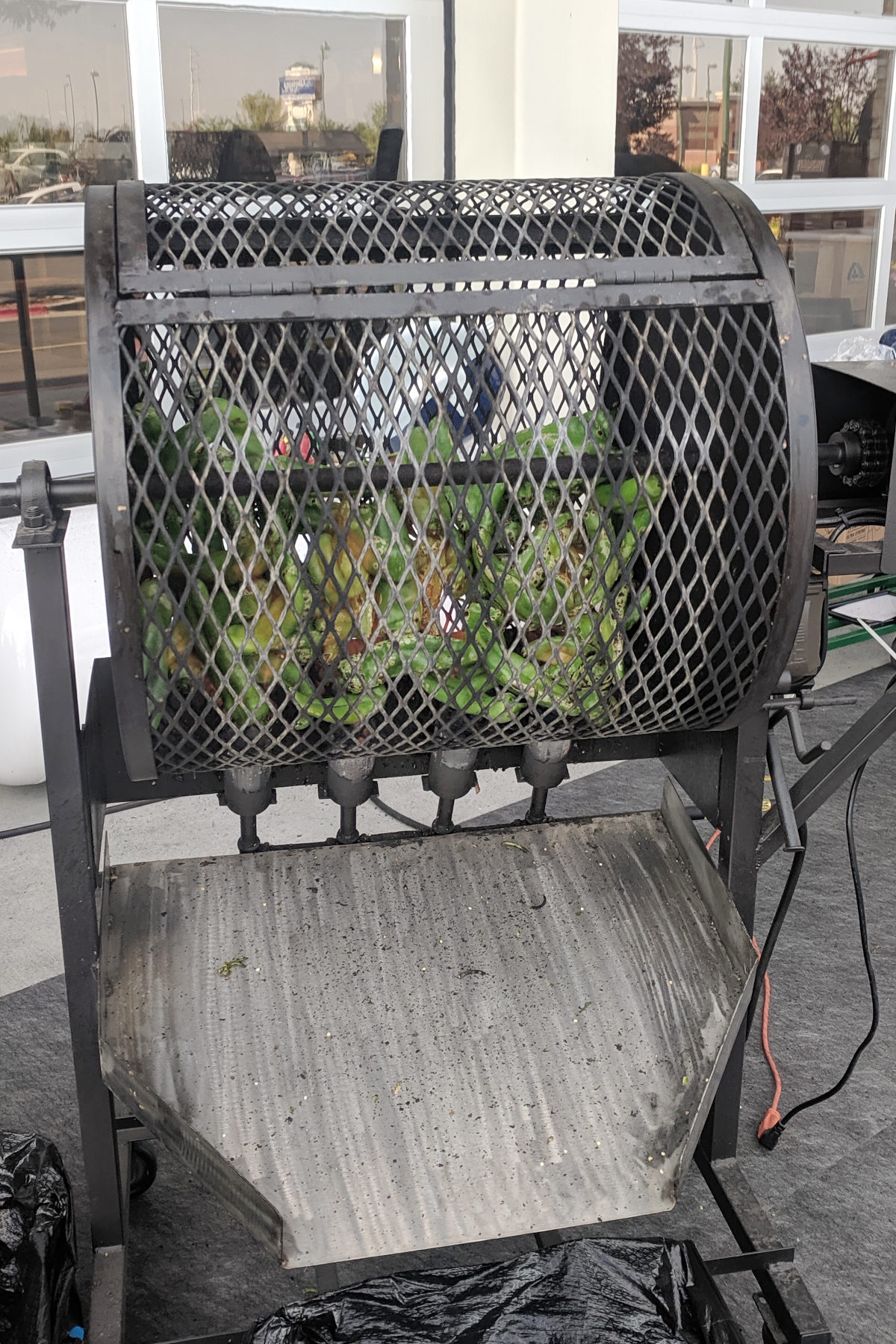 A metal roasting wheel drum filled with fresh green hatch chiles, spinning over open flame outside a grocery store, with a metal catch tray underneath to collect the roasted peppers.