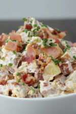 Loaded potato salad piled high in a white bowl, with crispy bacon pieces, red potato chunks, and fresh chives mixed into a creamy dressing.