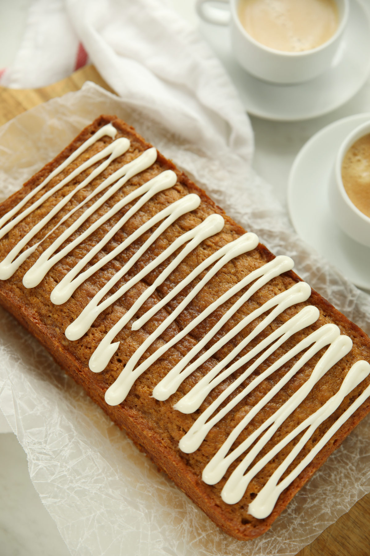 Loaf of pumpkin sourdough bread with cream cheese glaze on parchment paper, next to two cups of coffee.
