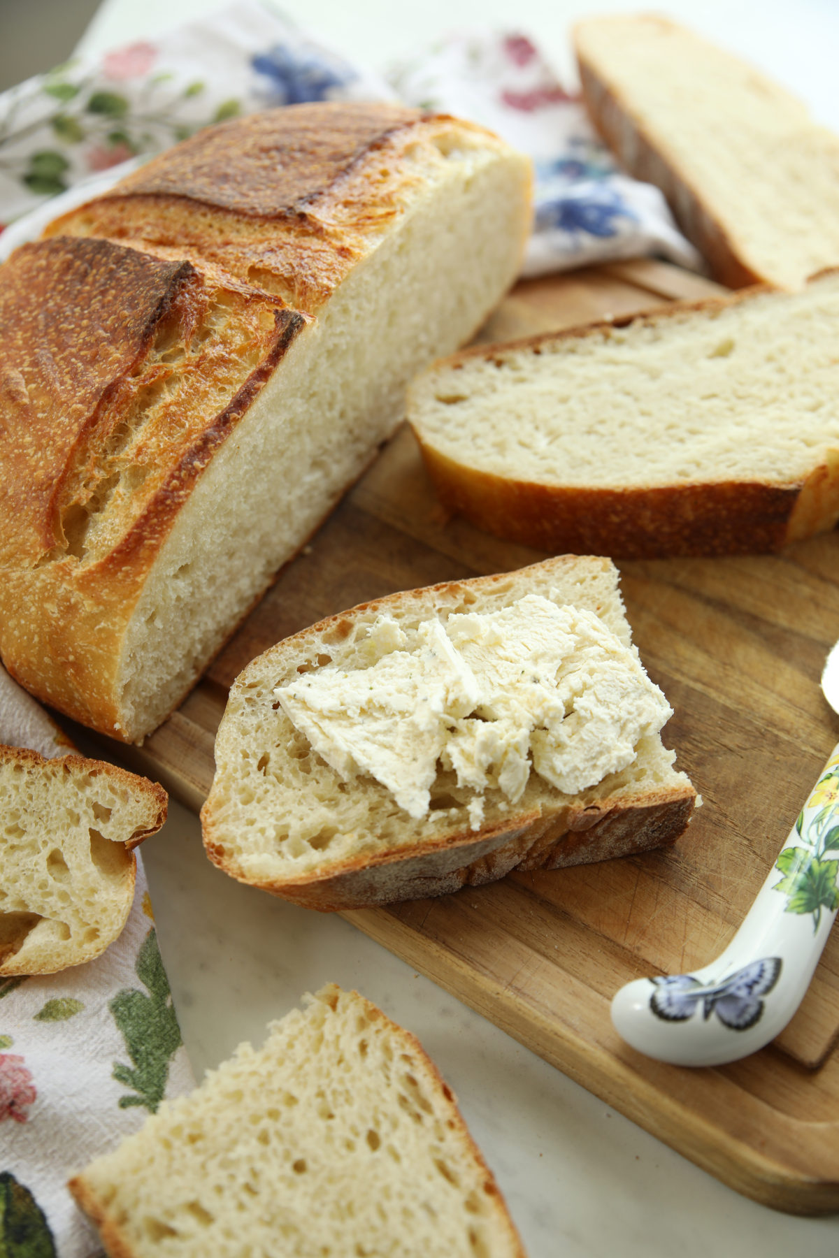 A loaf of protein sourdough bread sliced open on a wooden cutting board, showing a soft, airy crumb, with one slice spread with butter next to a floral tea towel