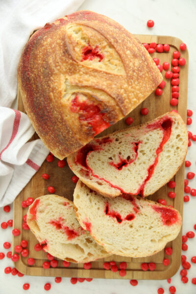 Alt text: "Sliced Red Hot Cinnamon Sourdough Bread showing red candy swirled through the crumb, on a wood board surrounded by scattered red hot candies.