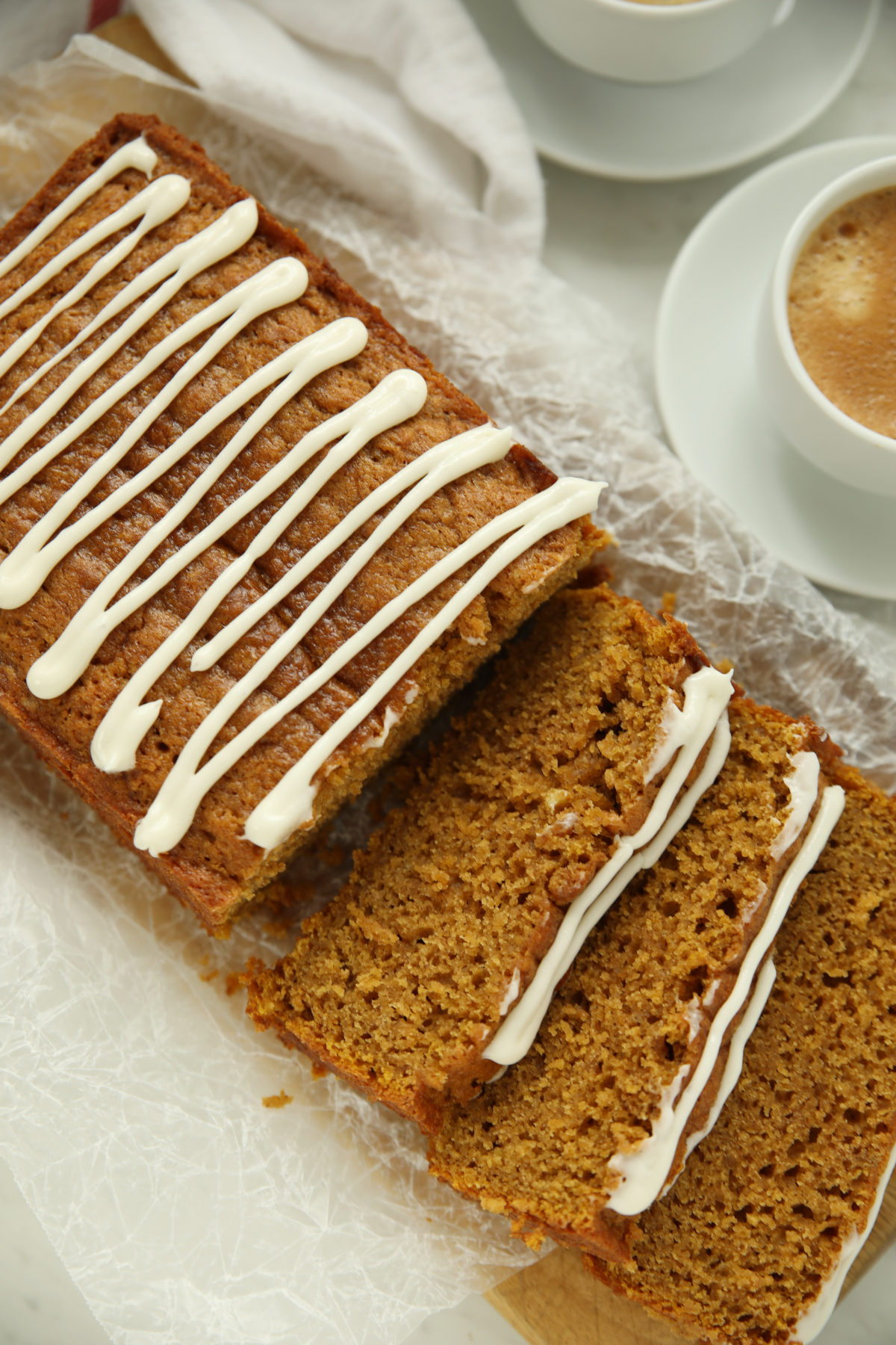 Sliced pumpkin sourdough bread with cream cheese glaze on parchment paper, next to two cups of coffee.